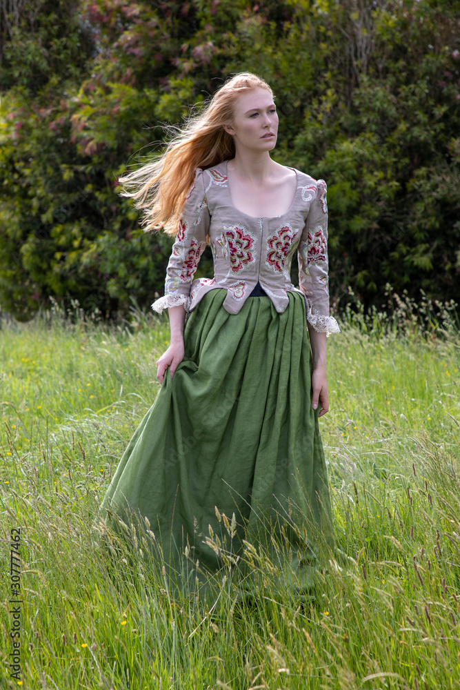 Red-haired 18th century woman in embroidered bodice and standing in long grass in the country side