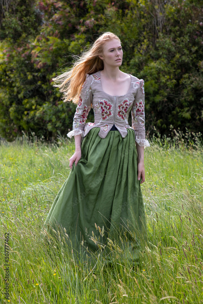 Red-haired 18th century woman in embroidered bodice and standing in long grass in the country side Red-haired 18th century woman in embroidered bodice and standing in long grass in the country side