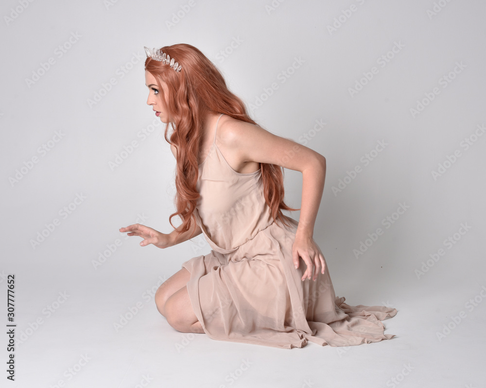 Fototapeta premium full length portrait of a pretty, fairy girl wearing a nude flowy dress and crystal crown. Seated pose against a grey studio background.