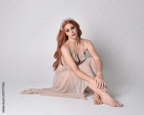 full length portrait of a pretty, fairy girl wearing a nude flowy dress and crystal crown. Seated pose against a grey studio background.