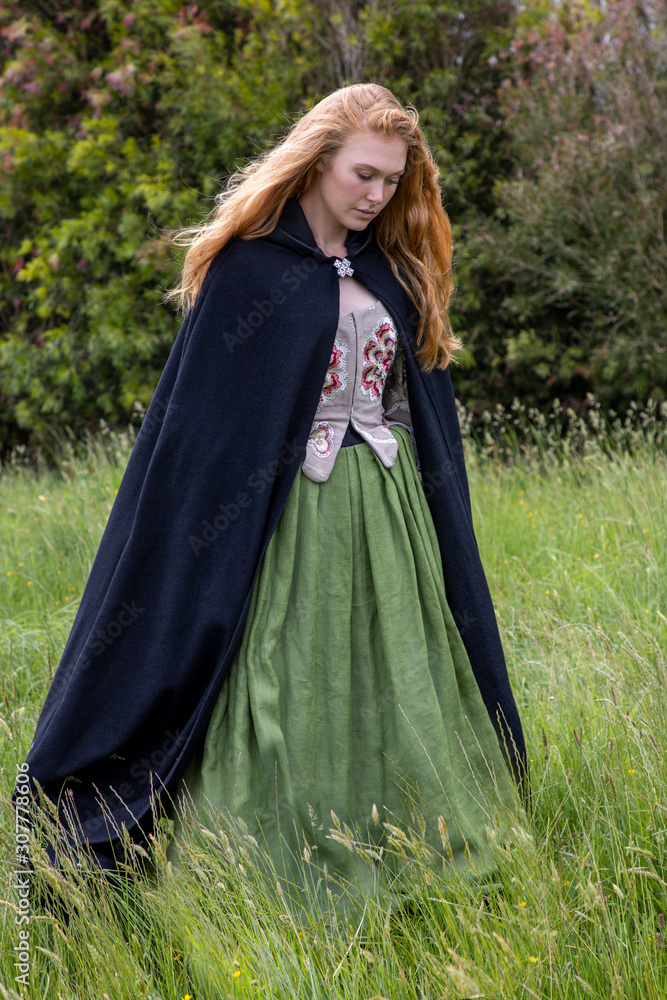 Red-haired 18th century woman in embroidered bodice and standing in long grass in the country side