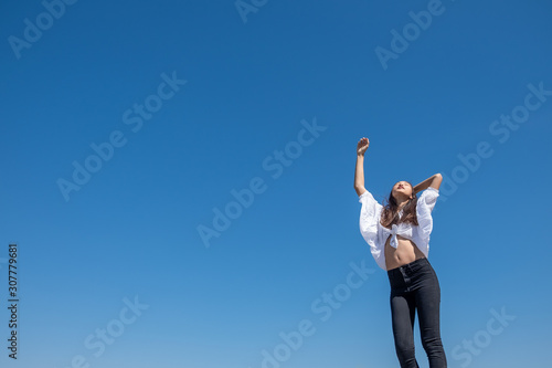 Happy woman with tanned slim body breathing fresh air raising her arms up, enjoying a sunny summer holiday on beach destination against blue sky, outdoors. Travel and well being lifestyle.