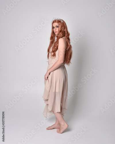 full length portrait of a pretty, fairy girl wearing a nude flowy dress and crystal crown. Standing and dancing pose against a grey studio background.