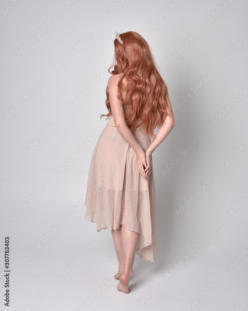 Fototapeta premium full length portrait of a pretty, fairy girl wearing a nude flowy dress and crystal crown. Standing and dancing pose against a grey studio background.
