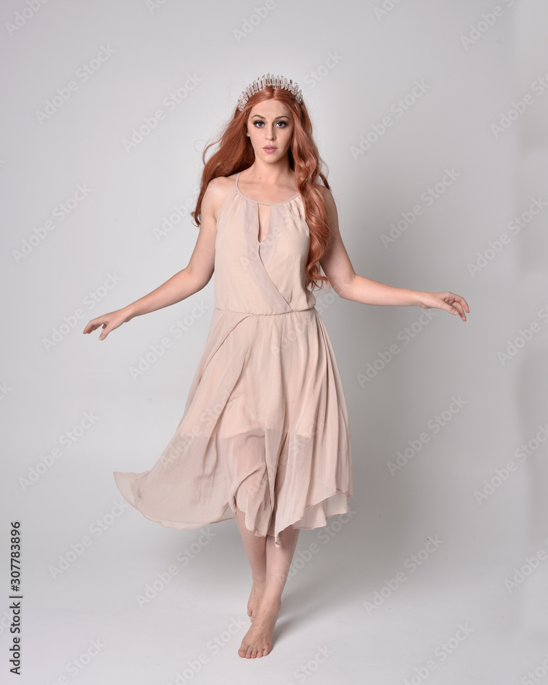 full length portrait of a pretty, fairy girl wearing a nude flowy dress and crystal crown. Standing and dancing pose against a grey studio background.
