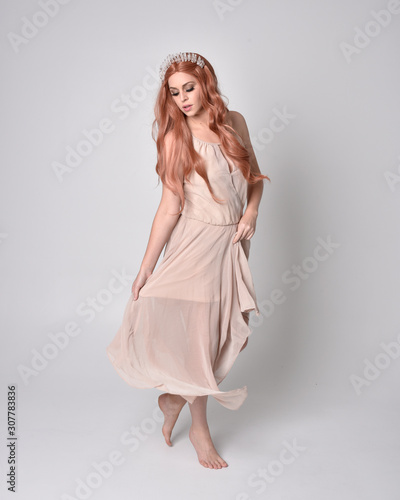 full length portrait of a pretty, fairy girl wearing a nude flowy dress and crystal crown. Standing and dancing pose against a grey studio background.