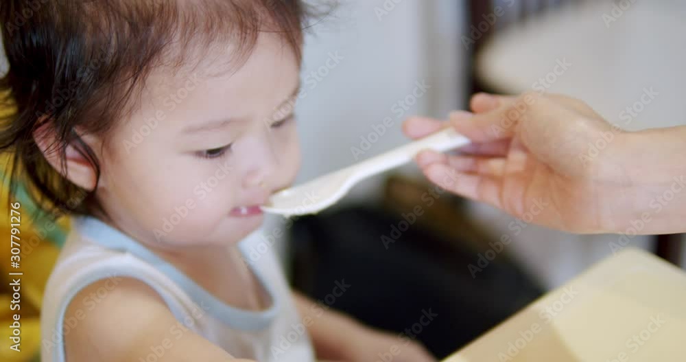 Asian Grandmother using spoon feeding food to her daugther.Cute baby girl enjoy eating lunch while sitting on her chair in livingroom at home.love and care concepts. Asian Grandmother using spoon feeding food to her daugther.Cute baby girl enjoy eating lunch while sitting on her chair in livingroom at home.love and care concepts.