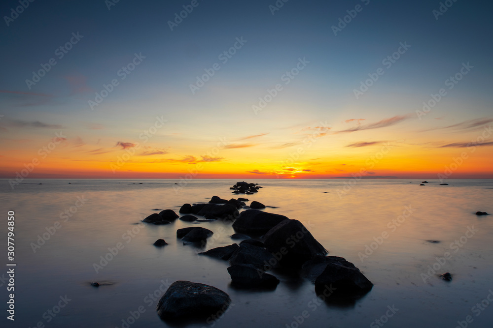 Vibrant colored summer sunset reflecting in ocean with endless horizon and deep blue ocean, silhouette of boulders laying in the foreground in shallow water at island of Gotland, Sweden