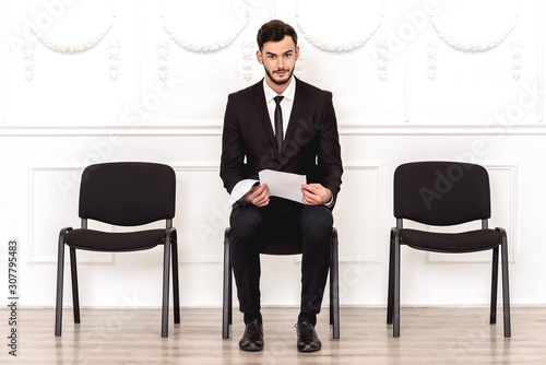 Job interview. Man sitting isolated on white waiting with resume looking camera pensive