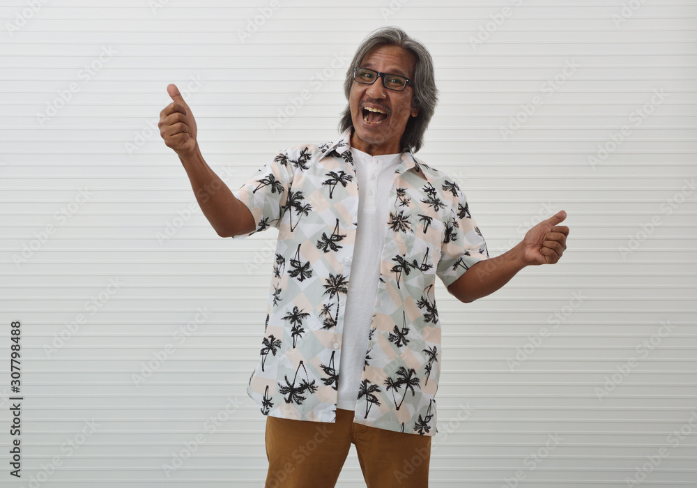 Naklejka premium Happy senior traveler asian man wearing glasses, summer shirt and brown shorts making thumbs up over white wall background, Business summer holiday concept