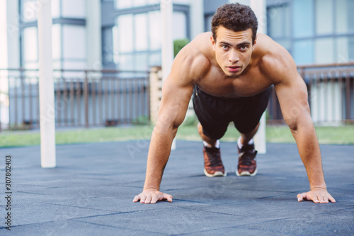 Strong muscular mixed race black man working out in outdoor gym sportsground. Crossfit training. Making push-ups.