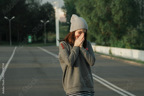  Portrait of a young woman who covers her face with her hands and laughs on the background of the road, in a gray hat with dark hair and a gray sweatshirt. Summer.