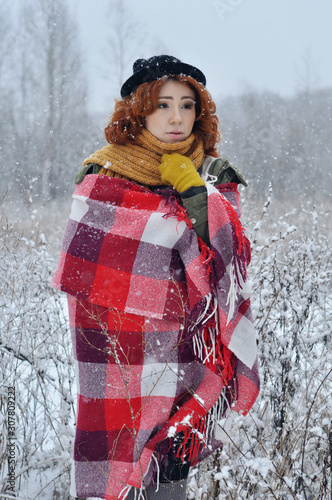 Cheerful red-haired girl warms her hands wrapped in a warm red checkered plaid during a snowfall