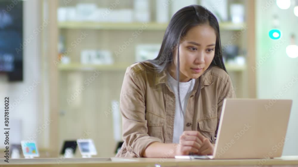 Medium shot of young Asian woman choosing laptop computer at gadget store and testing one of them Medium shot of young Asian woman choosing laptop computer at gadget store and testing one of them