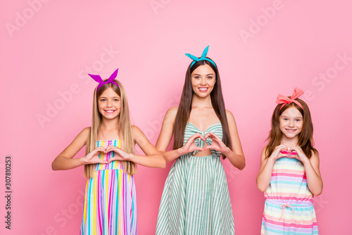Portrait of cheerful ladies kid making heart shaped signs with their fingers wearing headbands skirt dress isolated over pink background