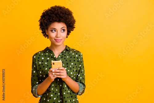 Photo of beautiful funny dark skin wavy lady hold telephone hands read good youth news look empty space advert wear green dotted shirt isolated yellow color background