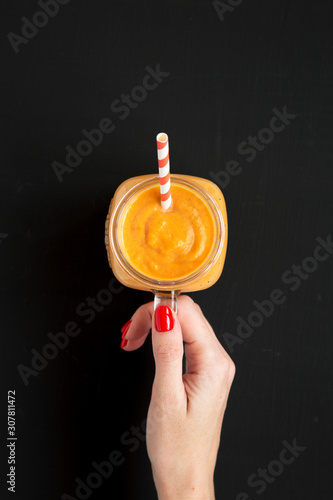 Female hand holds a glass jar with pumpkin smoothie over black background, top view. Flat lay, overhead, from above.