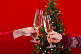 Cropped chot of couple in love clinking champage glasses, toasting next to Christmas tree, wearing themed sweaters. Man and woman celebrating new year. Close up, copy space, red wall background.