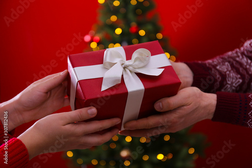 Cropped chot of couple in love exchanging the presents next to Christmas tree, wearing themed sweaters. Man and woman celebrating new year. Close up, copy space, red wall background. photo