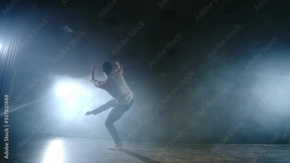 A modern ballet, a man performs jumps and spins in the light of spotlights and smoke on a dark background. Acrobatic choreography, rehearsal of the script of modern ballet A modern ballet, a man performs jumps and spins in the light of spotlights and smoke on a dark background. Acrobatic choreography, rehearsal of the script of modern ballet