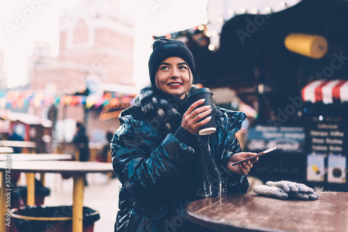 Modern happy woman with coffee cup using smartphone at Christmas market