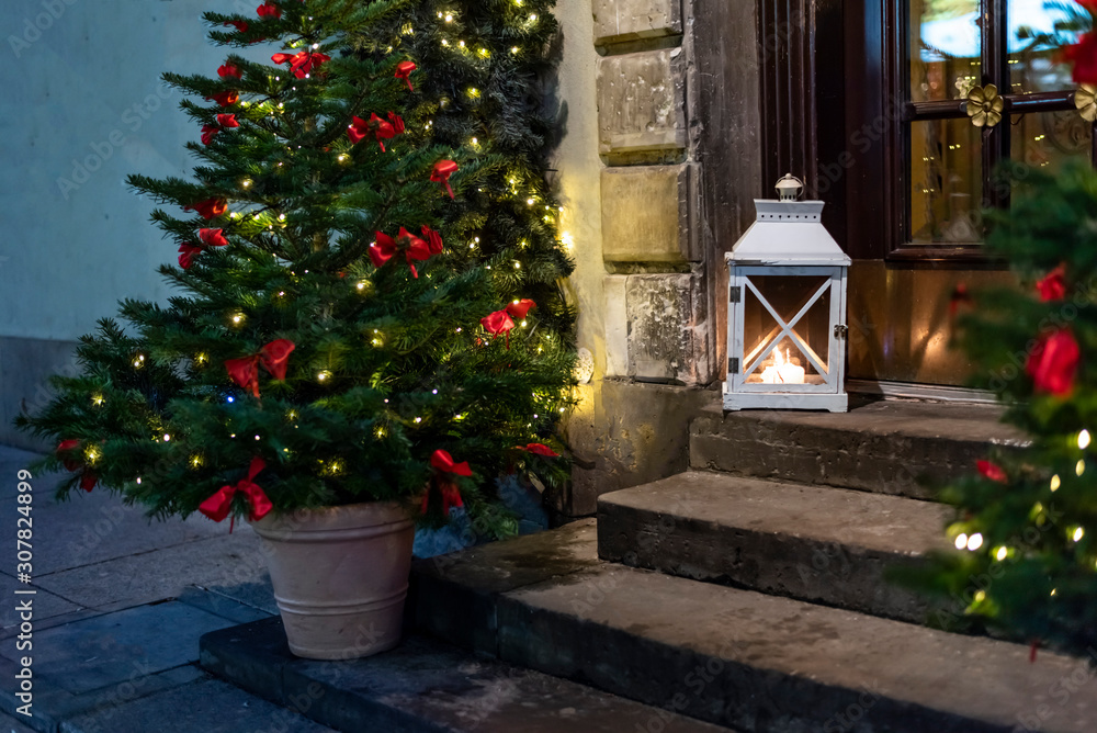 Christmas trees decorated with garland lightings and bowknots of red ribbon, vintage lantern outdoors on doorsteps in european old town house on christmas eve