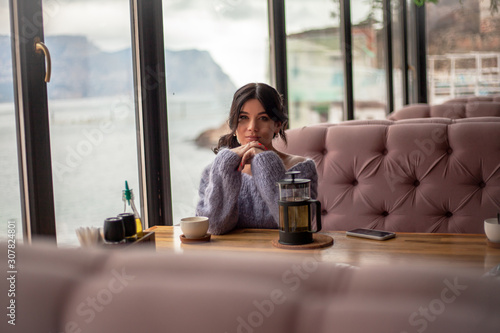 Young pregnant woman sit in cozy cafe with beautiful view of window of the sea and mountains drink healthy tea . Female dressed warm woolen sweater and siting on the sofa.