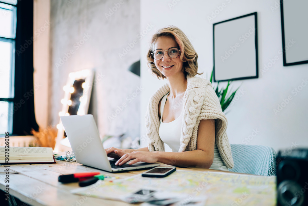 Obraz premium Portrait of cheerful hipster girl in classic optical glasses sitting at desktop in loft apartment and smiling at camera during free time for search funny comedy movies on social website, freelance