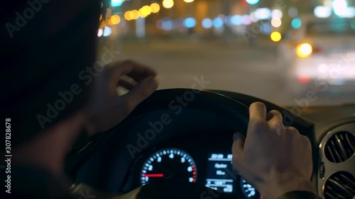 Close up man's hands on the steering wheel of a car. Dashboard lights up inside the dark interior of a car while driving at night. POV. Blurred lights on the city street