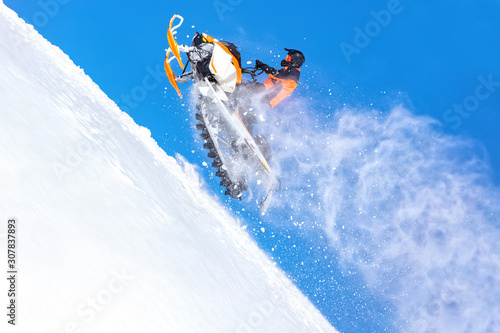 elite sports snowmobiler rides and jumps on steep mountain slope with swirls of snow storm. background of blue sky leaving a trail of splashes of white snow. bright snowmobile and suit without brands