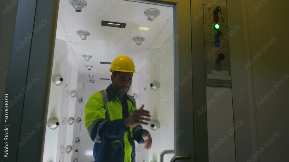 Black african worker in uniform and helmet opens door of air shower chamber. Remove dust and particles prepare to enter clean room. Jets blow air through filters. Foreman does protective procedures Black african worker in uniform and helmet opens door of air shower chamber. Remove dust and particles prepare to enter clean room. Jets blow air through filters. Foreman does protective procedures
