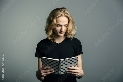 A young hipster guy with long blonde hair read notebook, . On a gray background.