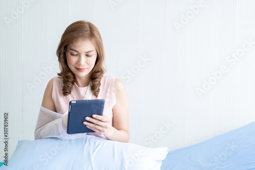 Asian woman broken arm sitting on the bed in hospital using a tablet. Concept of healthcare in modern lifestyle. Photograph with copy space.