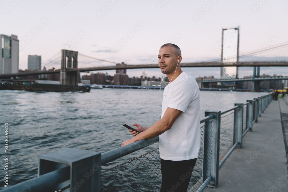 Ethnic short haired man with smartphone and earphones near river