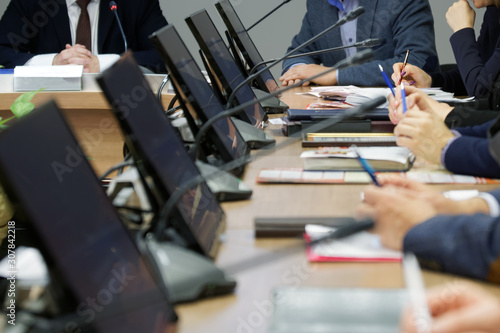 Men and women, businessmen or officials - participants in a meeting in a meeting room. A large table with installed monitors and microphones