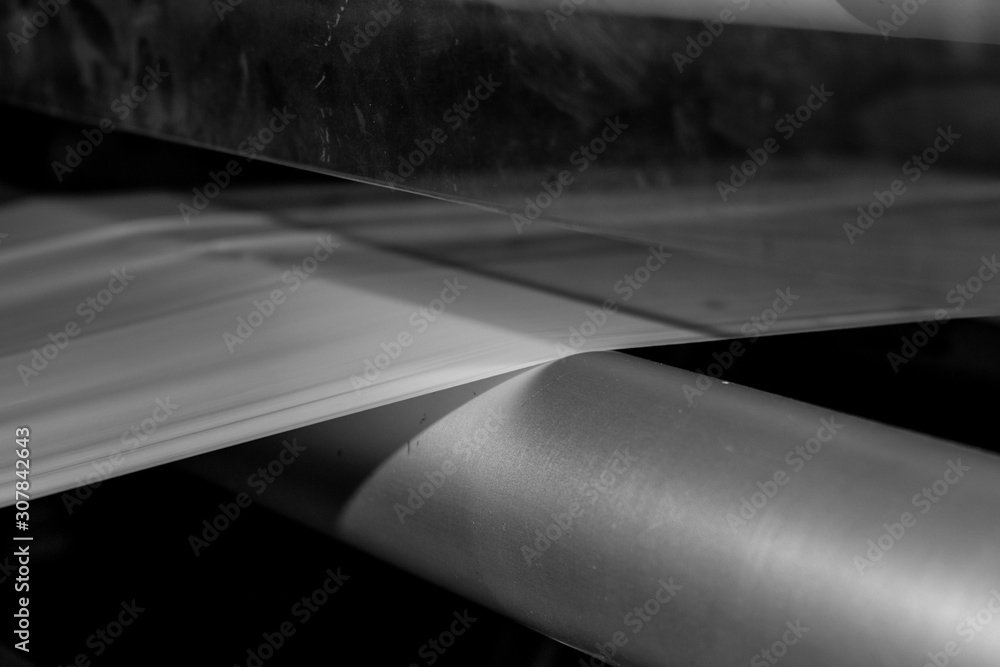 Black & White imagery Thin plastic sheet begin fed into a pressing cutting  machine via metal rollers with a reflection of the material in shiny metal  plate on factory cover. Stock Photo
