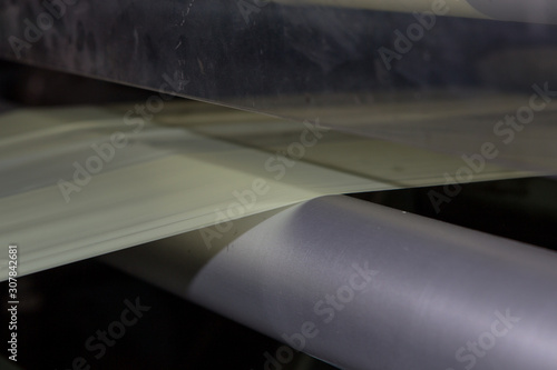 Imagery of light yellow Thin plastic sheet begin fed into a pressing cutting machine via metal rollers with a reflection of the material in shiny but dirty marked metal plate on factory cover.