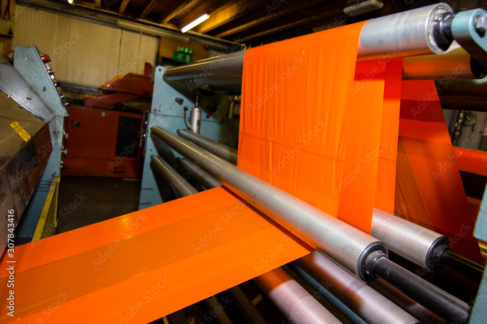 image of the inside workings of a machine with orange high vis plastic sheet being fed through industrial factory on rollers with tension keeping it straight being stretched and cut ready for packing