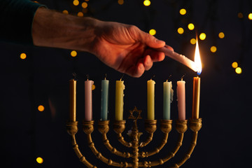 Partial view of man lighting up candles in menorah on black background with bokeh lights on Hanukkah