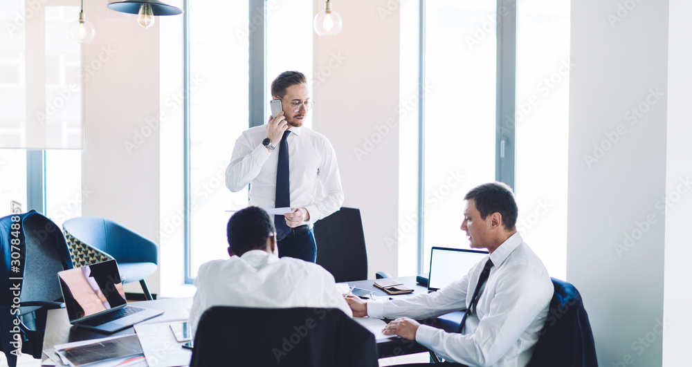 Adult businessman with papers standing and phoning while working with diverse employees in office Adult businessman with papers standing and phoning while working with diverse employees in office