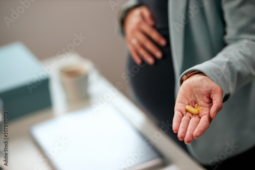 Close up of caucasian pregnant business woman standing in office and holding pills in hand.