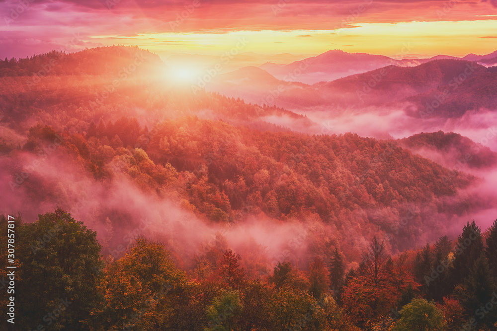 Fototapeta premium Amazing nature landscape, misty colorful sunrise in Alps, scenic view of wooded mountains with autumn trees, dramatic cloudy sky and rising sun. Outdoor travel background, Jamnik, Slovenia