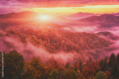 Amazing nature landscape, misty colorful sunrise in Alps, scenic view of wooded mountains with autumn trees, dramatic cloudy sky and rising sun. Outdoor travel background, Jamnik, Slovenia