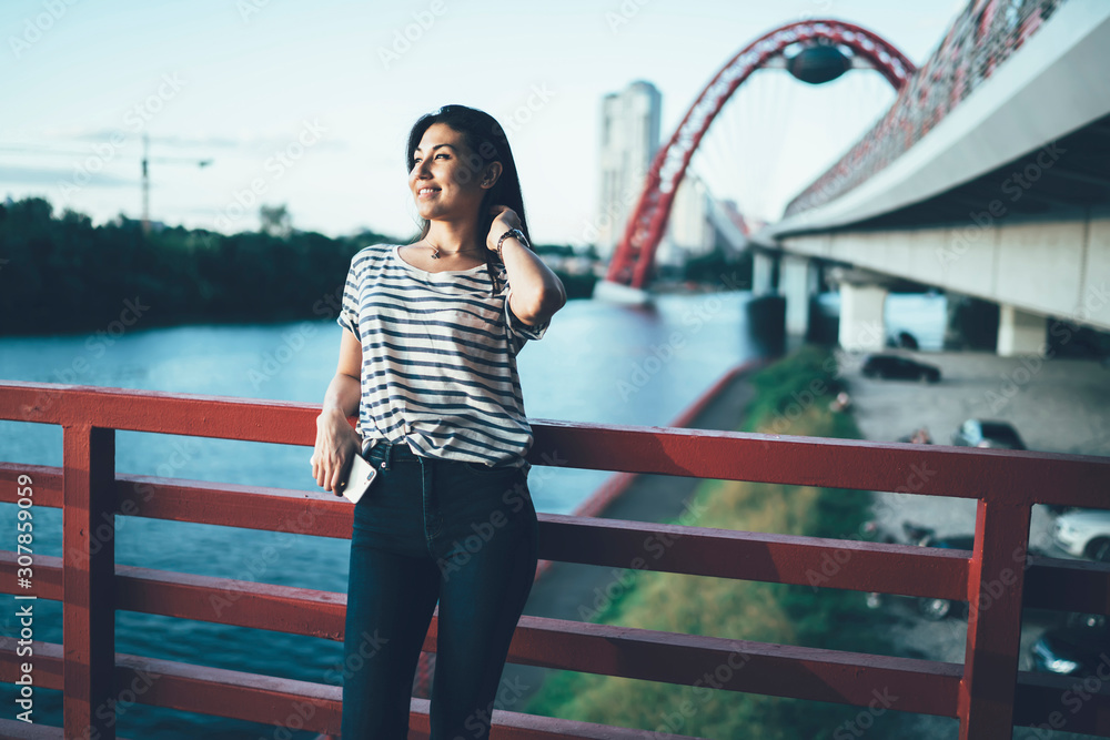 Asian hipster girl in casual wear recreating during leisure on city bridge during sunny summer day, smiling young woman traveler standing on urban setting background recreating on destination .