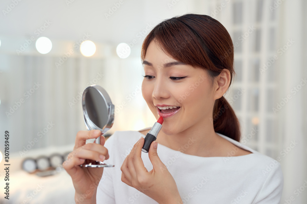  Smiling young Asian woman applying lipstick and looking to mirror.