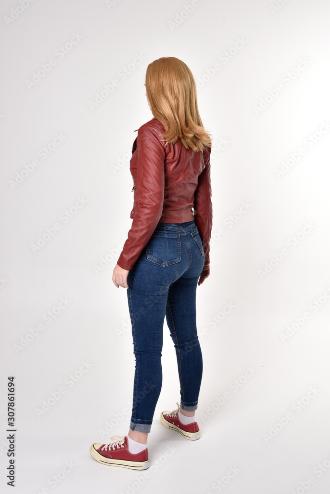 Fototapeta premium Full length portrait of a pretty blonde girl wearing red leather jacket denim jeans and sneakers. Standing pose, facing away from the camera, on a studio background.