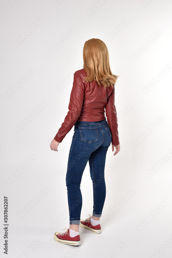 Full length portrait of a pretty blonde girl wearing red leather jacket denim jeans and sneakers. Standing pose, facing away from the camera, on a studio background. Full length portrait of a pretty blonde girl wearing red leather jacket denim jeans and sneakers. Standing pose, facing away from the camera, on a studio background.