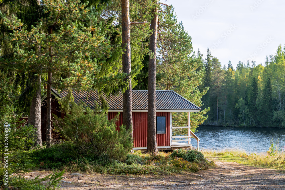 Red wooden finnish traditional cabins cottages in green pine forest near river. Rural architecture of northern Europe. Wooden houses in camping on sunny summer day