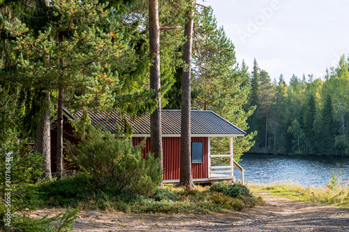 Red wooden finnish traditional cabins cottages in green pine forest near river. Rural architecture of northern Europe. Wooden houses in camping on sunny summer day
