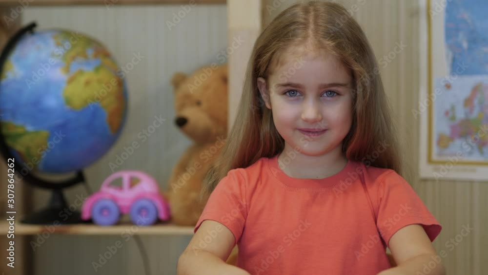 Close Up Portrait of Little Girl Looking Camera. Face Portrait Closeup. Pupil Little Kid Looking at Camera and Smiling Kindly. Cute Smart Positive Small Schoolgirl Sitting at Desk. Close Up Portrait of Little Girl Looking Camera. Face Portrait Closeup. Pupil Little Kid Looking at Camera and Smiling Kindly. Cute Smart Positive Small Schoolgirl Sitting at Desk.
